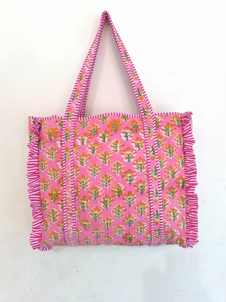 May include: Pink tote bag with a floral pattern in orange and white. The bag has matching pink handles and ruffled edges. The bag is made of a quilted fabric.