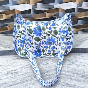 Puede incluir: Un bolso blanco con un estampado floral azul y hojas verdes. El bolso tiene un ribete a rayas azules y blancas y asas a juego. El bolso es de tela y está diseñado para llevarlo al hombro.