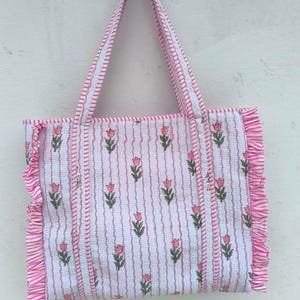 May include: A white tote bag with a pink floral pattern and vertical stripes. The bag has pink ruffled edges and matching pink handles. The floral design features pink tulips with green leaves. The bag is made of a soft, patterned fabric.