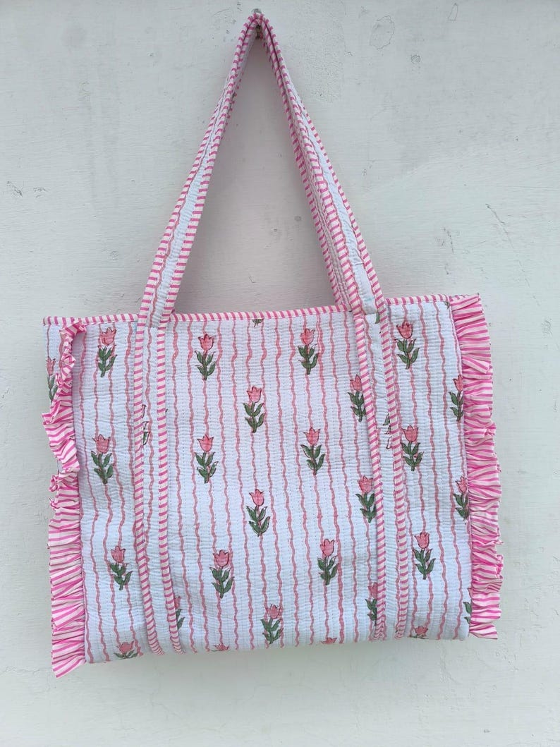 May include: A white tote bag with a pink floral pattern and vertical stripes. The bag has pink ruffled edges and matching pink handles. The floral design features pink tulips with green leaves. The bag is made of a soft, patterned fabric.