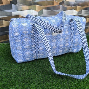 May include: A blue and white floral patterned duffel bag with striped handles. The bag is made of quilted fabric and has a rectangular shape. The background features a textured wall and green grass.