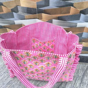 May include: Pink tote bag with a floral pattern and striped accents. The bag features a matching interior pocket and handles. The bag has a quilted texture and ruffled edges. The bag is made of a soft fabric.