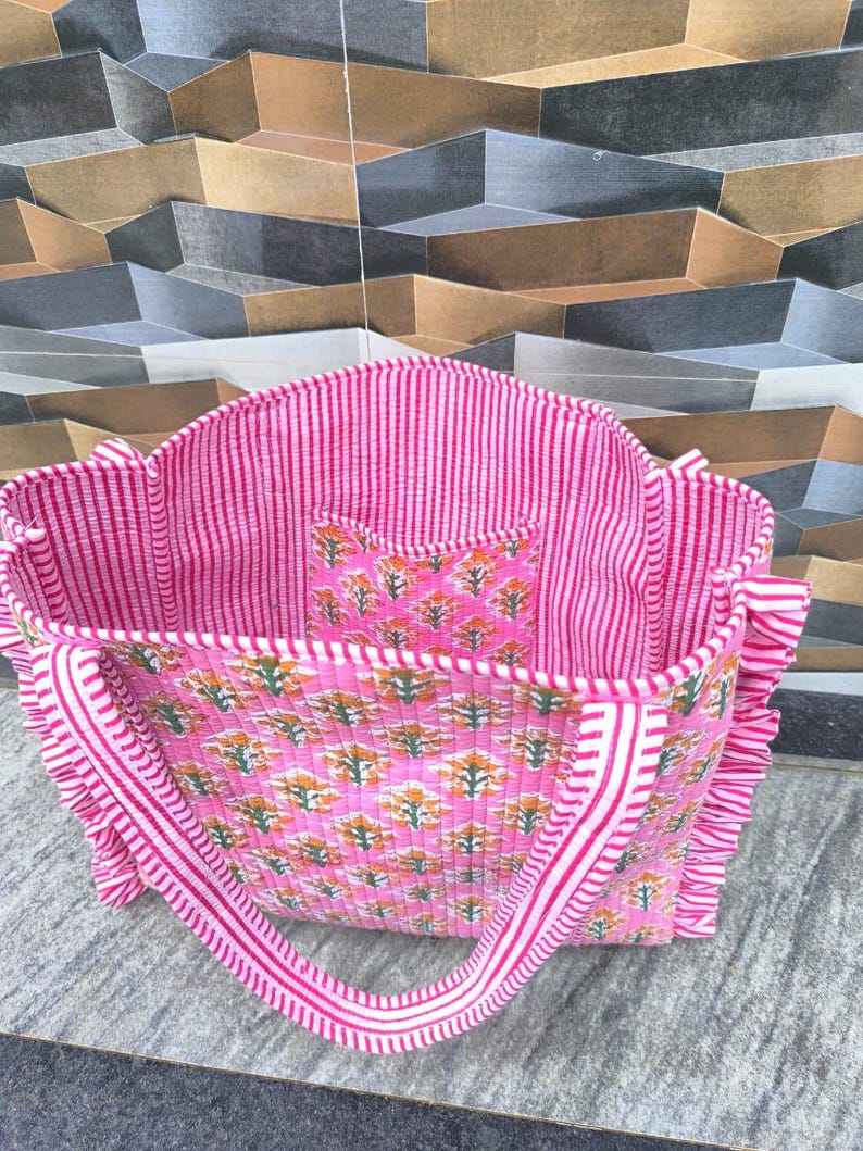 May include: Pink tote bag with a floral pattern and striped accents. The bag features a matching interior pocket and handles. The bag has a quilted texture and ruffled edges. The bag is made of a soft fabric.