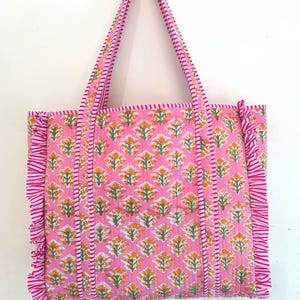 May include: Pink tote bag with a floral pattern in yellow and green. The bag has matching pink handles and ruffled edges. The floral pattern is repeated across the bag's surface, creating a cohesive design.
