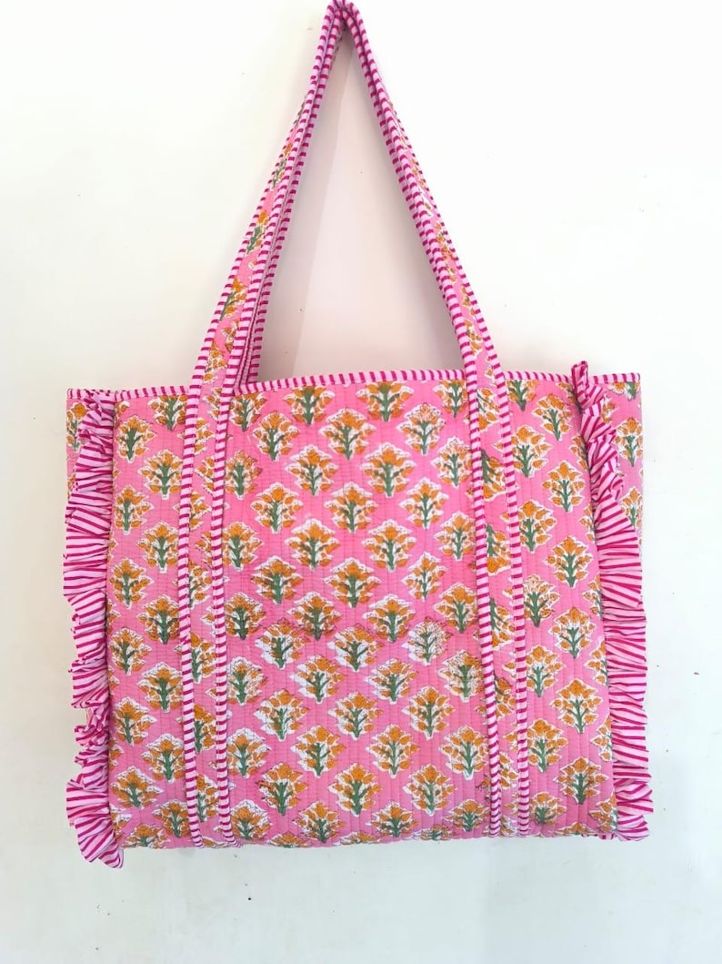 May include: Pink tote bag with a floral pattern in yellow and green. The bag has matching pink handles and ruffled edges. The floral pattern is repeated across the bag's surface, creating a cohesive design.