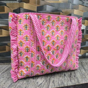 May include: Pink tote bag with a floral pattern in orange and green. The bag has a quilted design, ruffled edges, and striped handles. The bag is made of a soft, lightweight material.