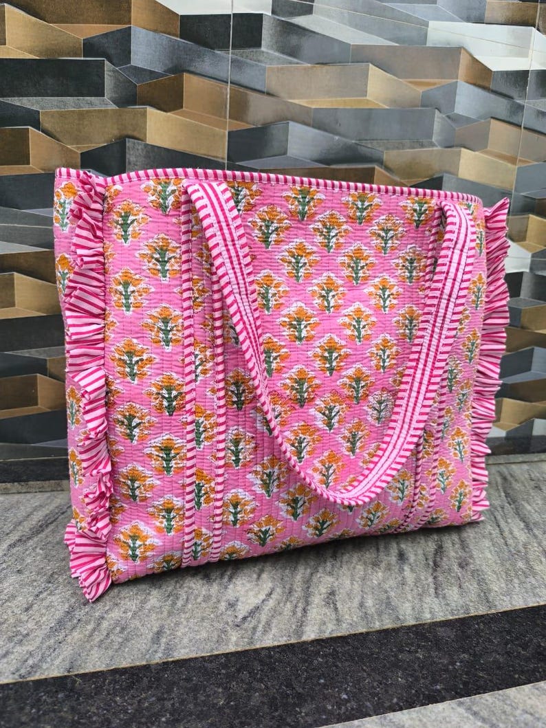 May include: Pink tote bag with a floral pattern in orange and green. The bag has a quilted design, ruffled edges, and striped handles. The bag is made of a soft, lightweight material.