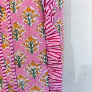May include: Pink floral patterned fabric with yellow flowers and green leaves. The fabric has a quilted design and a striped ruffle trim in shades of pink and white. The design is repeated across the fabric.