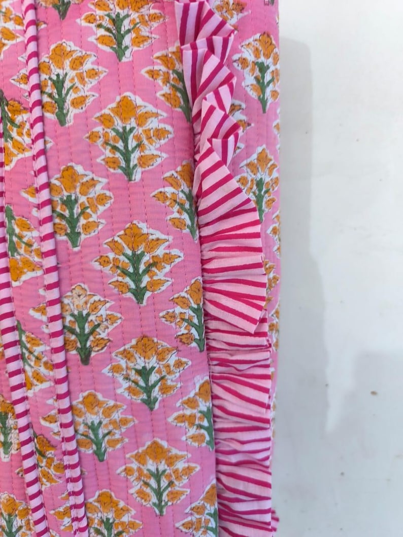 May include: Pink floral patterned fabric with yellow flowers and green leaves. The fabric has a quilted design and a striped ruffle trim in shades of pink and white. The design is repeated across the fabric.