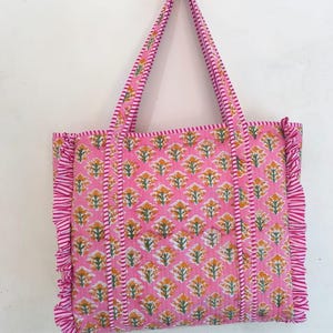 May include: Pink tote bag with a floral pattern in yellow and green. The bag has matching pink handles and ruffled edges. The floral pattern is repeated across the bag's surface, creating a cohesive design.
