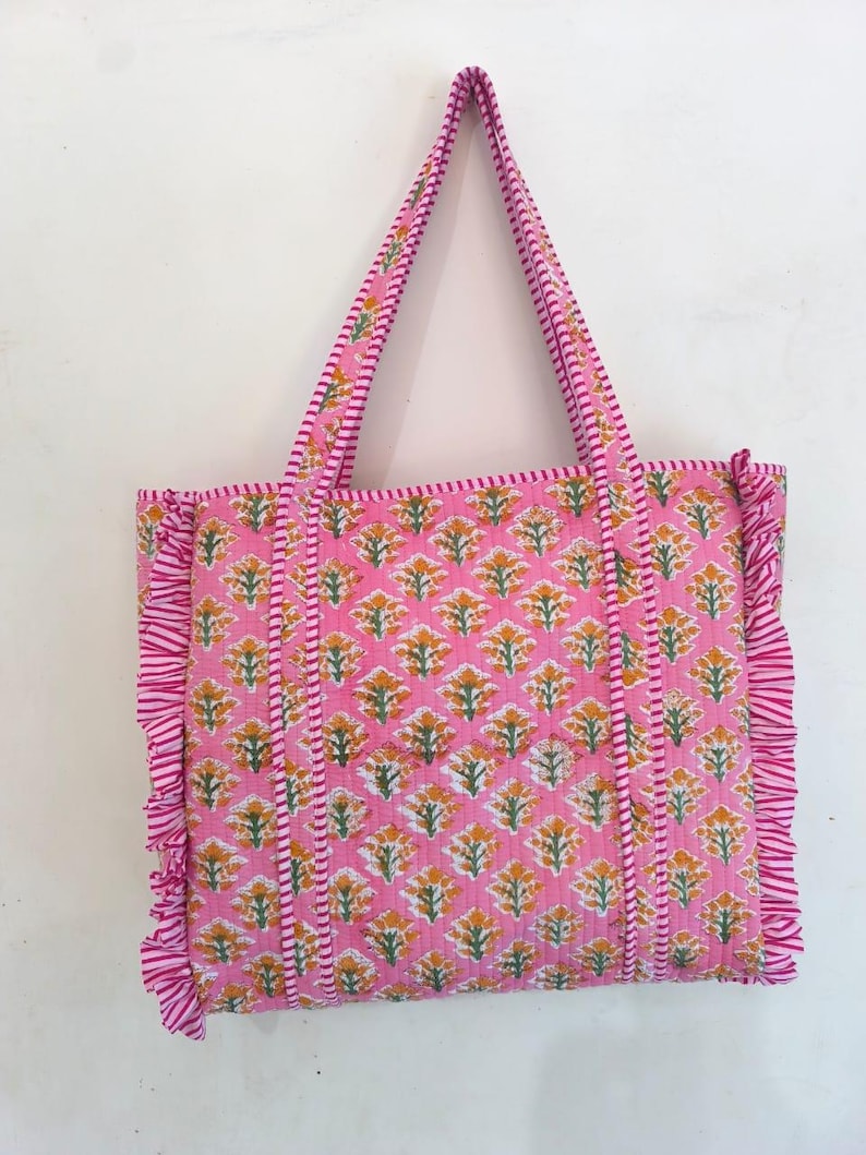 May include: Pink tote bag with a floral pattern in yellow and green. The bag has matching pink handles and ruffled edges. The floral pattern is repeated across the bag's surface, creating a cohesive design.