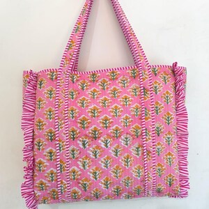 May include: A pink tote bag with a floral pattern in yellow and green. The bag has matching pink handles and ruffled edges. The floral pattern is repeated across the bag's surface. The bag is made of a quilted fabric.