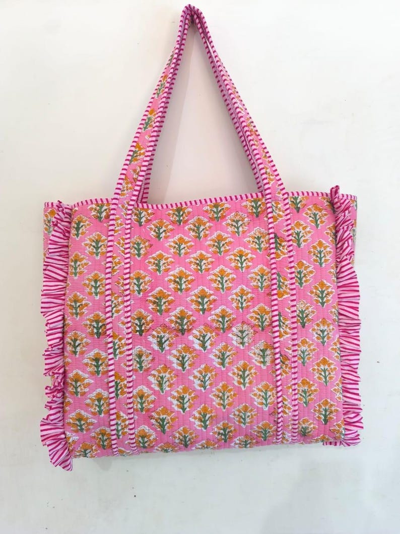 May include: A pink tote bag with a floral pattern in yellow and green. The bag has matching pink handles and ruffled edges. The floral pattern is repeated across the bag's surface. The bag is made of a quilted fabric.