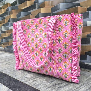 May include: Pink tote bag with a floral pattern in orange and green. The bag has a striped border and ruffled edges. The handles are also striped. The bag is made of a quilted fabric.