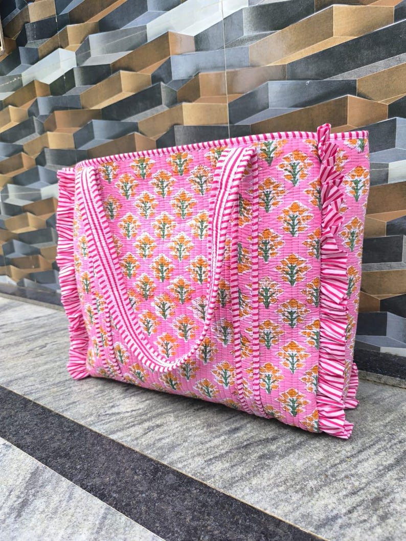 May include: Pink tote bag with a floral pattern in orange and green. The bag has a striped border and ruffled edges. The handles are also striped. The bag is made of a quilted fabric.