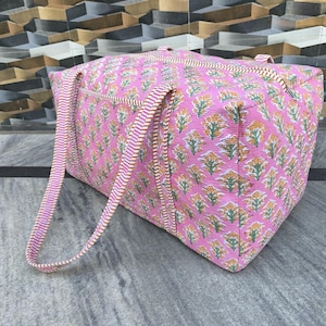 May include: A pink tote bag with a floral pattern in yellow and green. The bag has two handles with a striped design in pink and white. The bag is made of a quilted fabric.