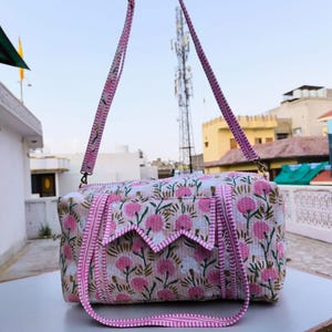 May include: A rectangular floral print handbag with pink and white flowers and green leaves. The bag has a pink strap and trim. The bag is sitting on a white surface.