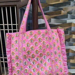 May include: Pink tote bag with a floral pattern in orange and green. The bag has a quilted design with ruffled edges and matching handles. The bag is hanging on a wooden post.
