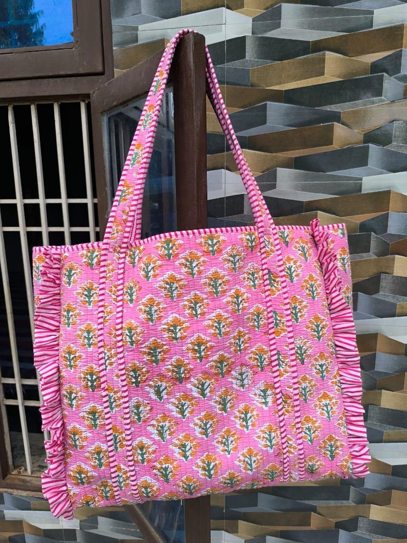 May include: Pink tote bag with a floral pattern in orange and green. The bag has a quilted design with ruffled edges and matching handles. The bag is hanging on a wooden post.