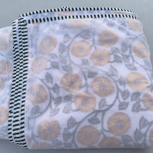 May include: Folded white fabric with a floral pattern in shades of gold and gray. The edges are trimmed with a black and white striped border. The fabric appears to be cotton or a similar lightweight material, suitable for bedding or home decor.