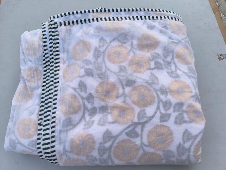 May include: Folded white fabric with a floral pattern in shades of gold and gray. The edges are trimmed with a black and white striped border. The fabric appears to be cotton or a similar lightweight material, suitable for bedding or home decor.