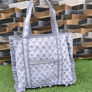 May include: White tote bag with a blue floral pattern and a zippered pocket. The bag has black and white striped handles and trim, and a ruffled edge. The bag is sitting on green grass.