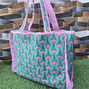 May include: A quilted tote bag with a floral pattern. The bag features a teal background with pink flower and leaf designs. The straps and trim are pink and white striped. The bag is sitting on green grass.