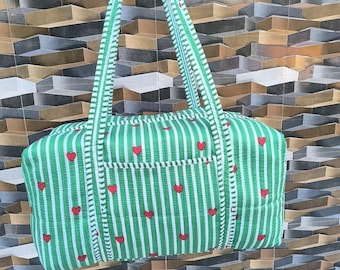May include: Green and white striped tote bag with red heart accents. The bag has a rectangular shape with a small pocket and two long handles. The bag is held up by a hand.