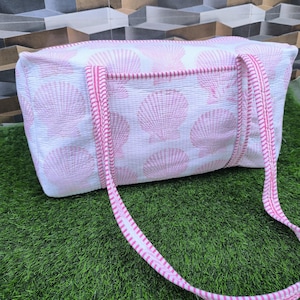 Puede incluir: Un bolso tote blanco y rosa con estampado de conchas marinas y asas rosas. El bolso es rectangular con cierre de cremallera y está hecho de tela acolchada. Las asas tienen un diseño a rayas rosas y blancas.