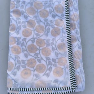 May include: White fabric with a floral pattern featuring beige flowers and gray leaves. The fabric has a border with black and white stripes. The fabric is folded over, showing the pattern and border design.