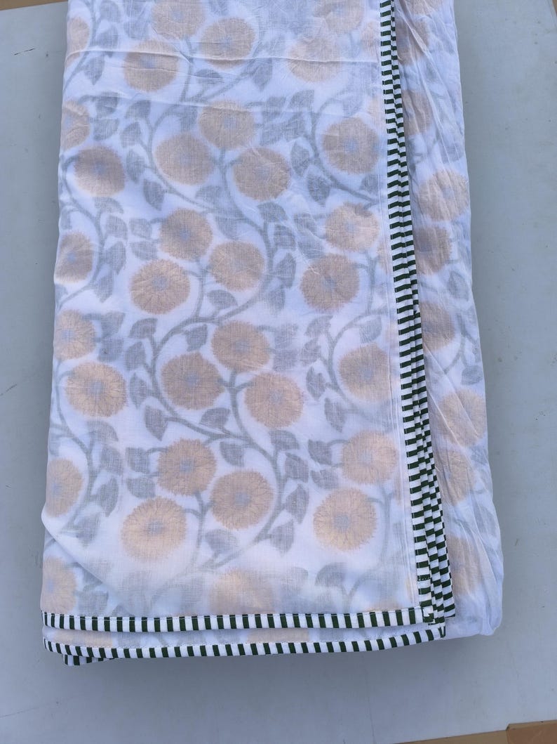 May include: White fabric with a floral pattern featuring beige flowers and gray leaves. The fabric has a border with black and white stripes. The fabric is folded over, showing the pattern and border design.