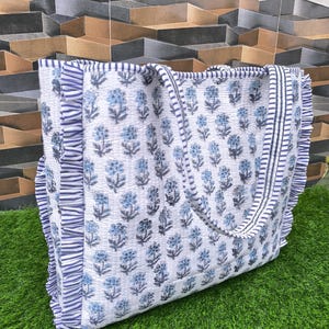 May include: A white tote bag featuring a blue floral design and frilled edges. The bag has blue and white striped trim and handles. The bag is on green grass.