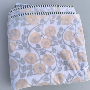 May include: Folded white fabric with a floral pattern featuring large, light orange flowers and gray leaves. The edges are trimmed with a green and white striped border. The fabric appears to be cotton or a similar material.