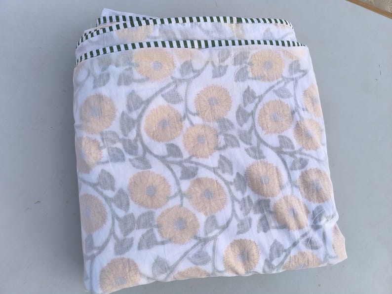 May include: Folded white fabric with a floral pattern featuring large, light orange flowers and gray leaves. The edges are trimmed with a green and white striped border. The fabric appears to be cotton or a similar material.