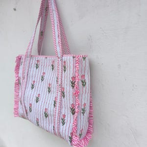 May include: A white tote bag with pink floral and stripe patterns. The bag features pink ruffled edges and matching handles. The floral design includes pink tulips and green leaves. The bag is hanging against a white wall.