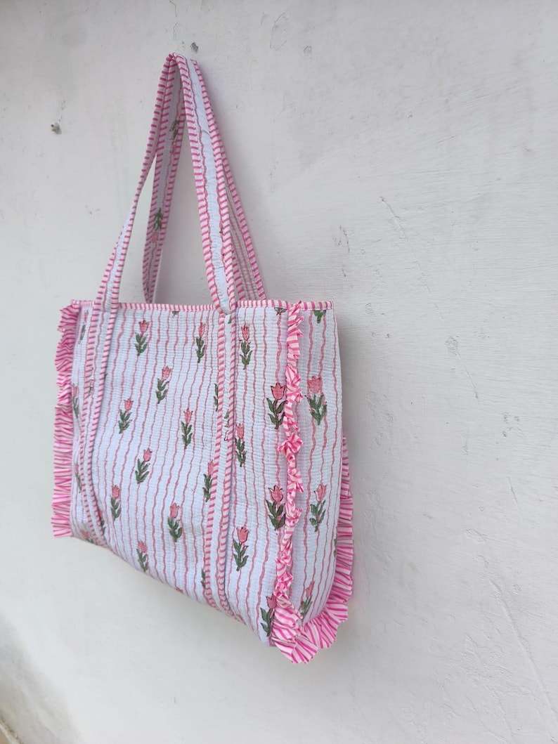 May include: A white tote bag with pink floral and stripe patterns. The bag features pink ruffled edges and matching handles. The floral design includes pink tulips and green leaves. The bag is hanging against a white wall.