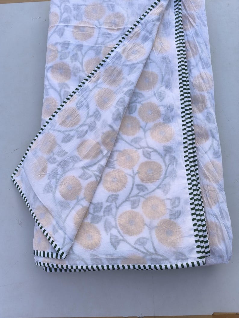 May include: White floral patterned fabric with a green and black checkered border. The fabric features a repeating pattern of yellow flowers and gray leaves. The fabric is folded, showing the pattern and border details.