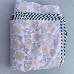 May include: White fabric with a floral pattern in yellow and gray. The fabric is folded, revealing a border with black and green stripes. The floral design features stylized flowers and leaves, creating a soft, natural aesthetic.