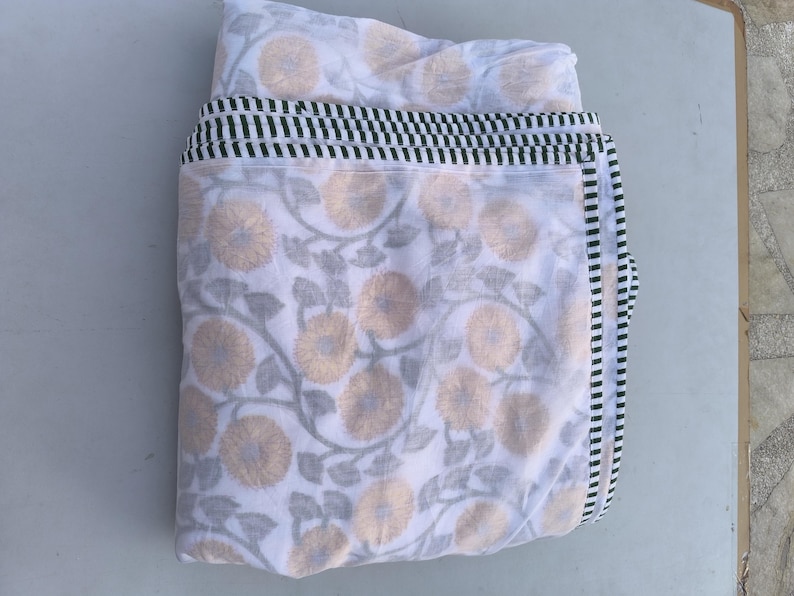 May include: White fabric with a floral pattern in yellow and gray. The fabric is folded, revealing a border with black and green stripes. The floral design features stylized flowers and leaves, creating a soft, natural aesthetic.