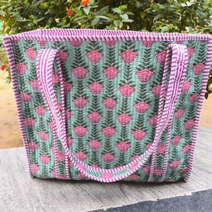 May include: A quilted tote bag with a floral pattern. The bag features a light green background with pink flower and leaf designs. The edges and handles are trimmed with pink and white striped fabric. The bag is sitting on a gray surface.