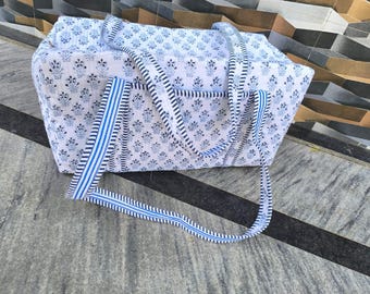 May include: A white tote bag with a blue floral pattern and blue and white striped handles. The bag is rectangular with a soft, padded appearance. The handles are long enough to be carried over the shoulder.