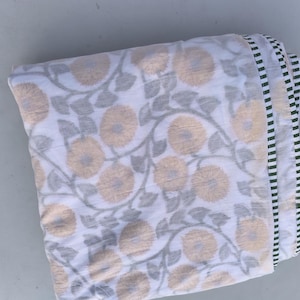 May include: Folded white fabric with a floral pattern in shades of beige and gray. The design features large, stylized flowers and leaves. The edge has a striped border in white, black, and green. This textile is suitable for home decor.