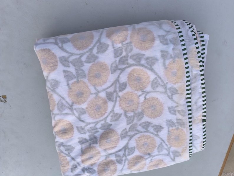 May include: Folded white fabric with a floral pattern in shades of beige and gray. The design features large, stylized flowers and leaves. The edge has a striped border in white, black, and green. This textile is suitable for home decor.