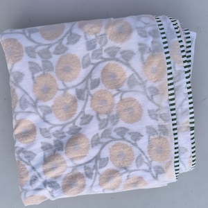 May include: Folded white fabric with a repeating pattern of large, light yellow flowers and gray leaves. The edge has a striped border in black, white, and green. The fabric appears to be a blanket or throw.
