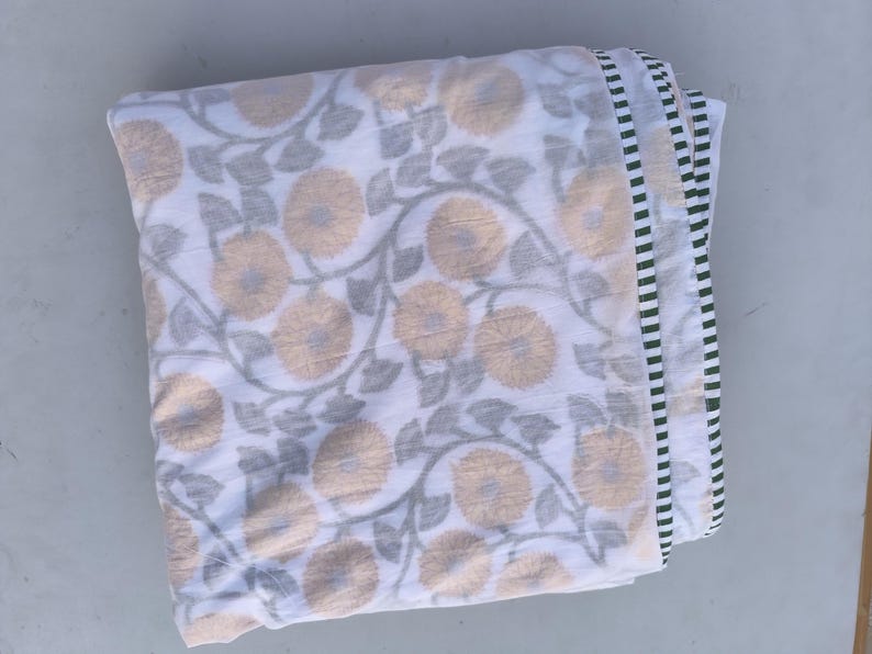 May include: Folded white fabric with a repeating pattern of large, light yellow flowers and gray leaves. The edge has a striped border in black, white, and green. The fabric appears to be a blanket or throw.