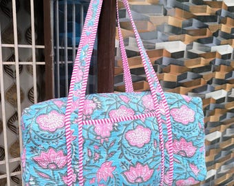 May include: A blue tote bag with a floral pattern in pink and white. The bag has pink straps and trim. The bag is made of a quilted fabric and has a pocket on the front.