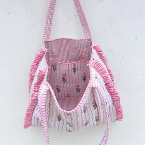 May include: A pink and white striped tote bag with a floral pattern. The bag has a rectangular shape with a ruffled edge and long straps. The interior and a small pocket feature a pink and white striped fabric with a floral design.