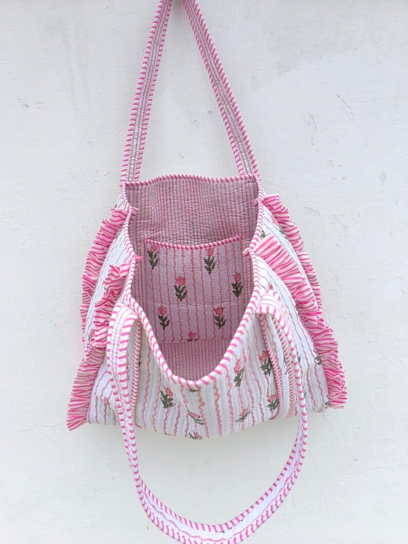 May include: A pink and white striped tote bag with a floral pattern. The bag has a rectangular shape with a ruffled edge and long straps. The interior and a small pocket feature a pink and white striped fabric with a floral design.