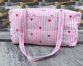 May include: Pink and white striped tote bag with red heart accents. The bag features a rectangular shape, a front pocket, and two handles. The bag is made of a quilted fabric.