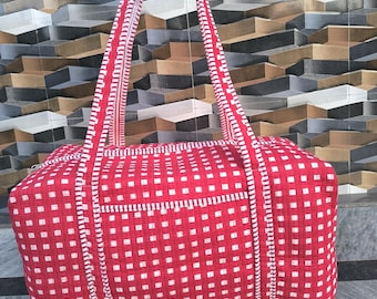 May include: A red and white patterned tote bag with long handles. The bag features a repeating square pattern and white stitching details. The handles are also red with white accents. The bag is held up by a hand.